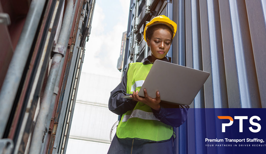 An African American female is standing between shipping containers and making notes on a laptop